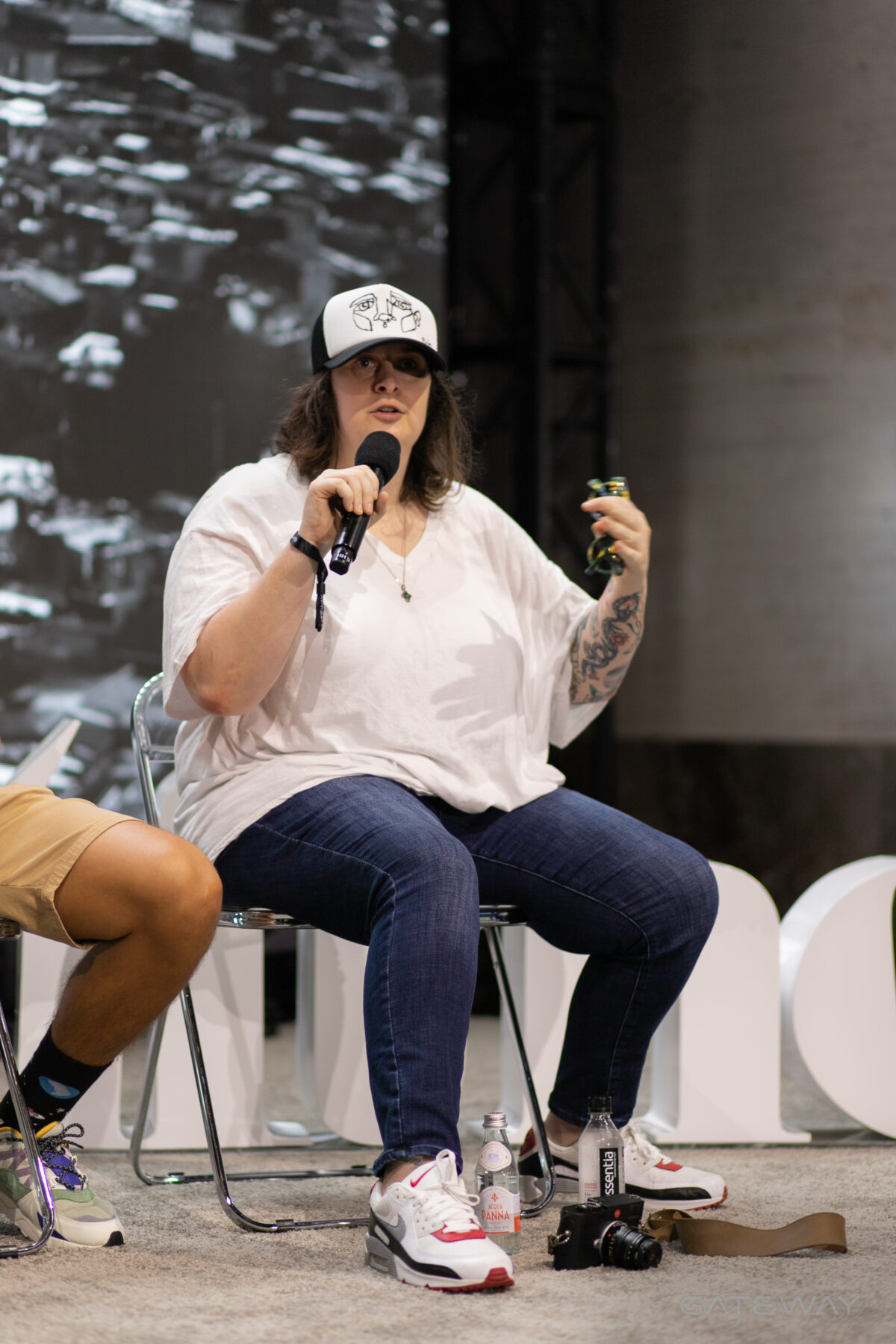 Une femme portant une casquette de baseball, une chemise blanche et un jean tient un microphone sur une scène et fait des gestes avec sa main tout en parlant. 