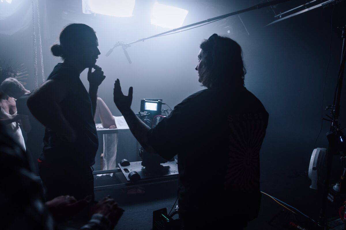 Une scène du tournage d'un film, de fortes lumières blanches au premier plan et la silhouette d'une femme et d'un homme qui font des gestes et se parlent.