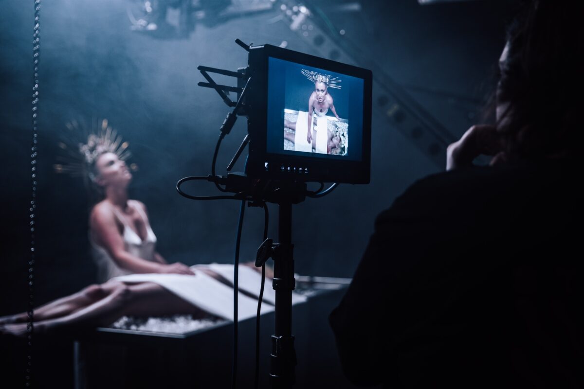 Un plateau de tournage sombre, une femme est derrière un moniteur regardant une scène au premier plan qui montre une femme vêtue d'une robe blanche et d'une étrange couronne tout en veillant sur un corps sur une table.